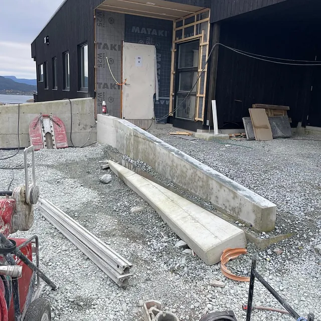Pågående bygging av et svart trehus med betongarbeid og bygningsmaterialer på bakken.