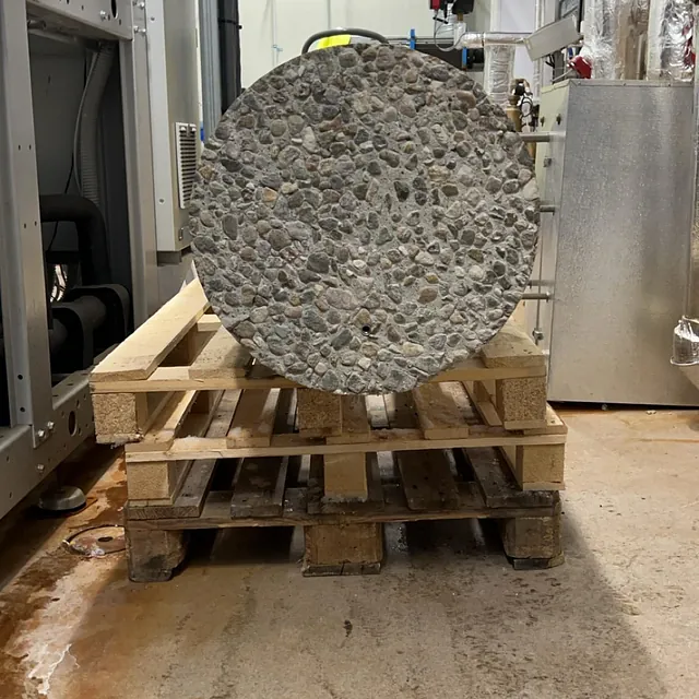 Stor betongsylinder med grov overflate ligger på trepaller i et teknisk rom.